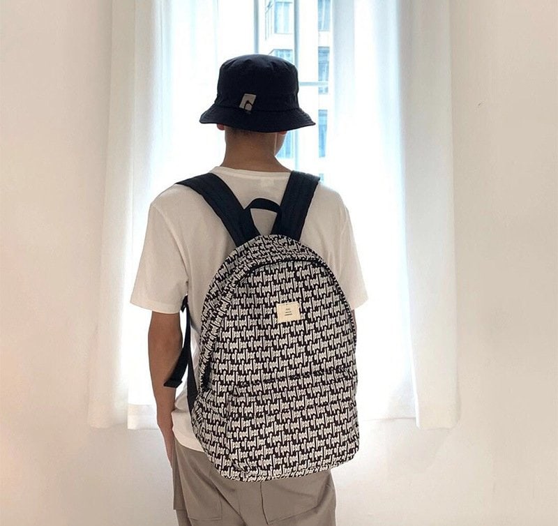 Backpack with black and white all-over print, typical of trendy kpop merchandise, worn by a person in casual attire, featuring a small logo patch at the top front.