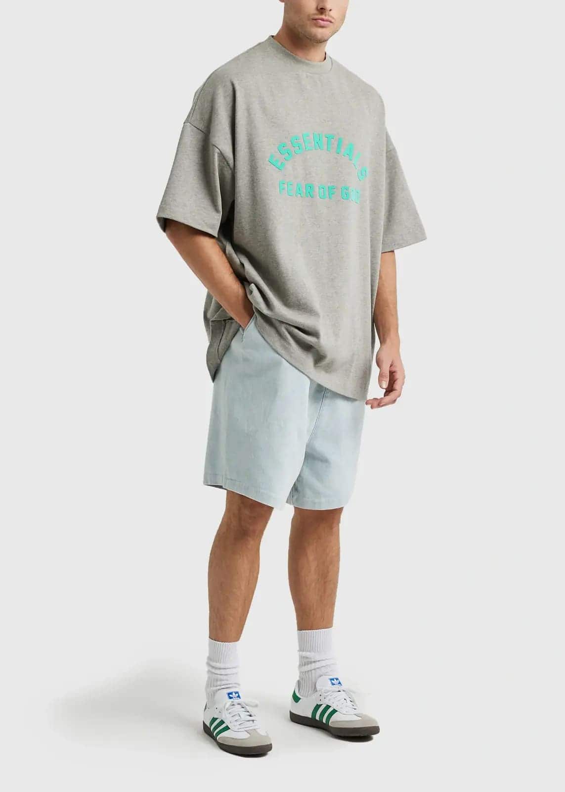 Comfy oversized grey T-shirt with blue graphic text "ESSENTIALS FEAR OF GOD", paired with light wash denim shorts, white sneakers with green accents, and white socks. Perfect for casual streetwear.
